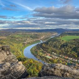 Panoramic view from Lilienstein rock formation showing the Elbe River winding through forested valleys toward Königstein under a cloudy sky.