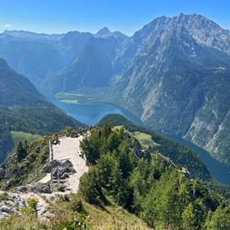 Panoramic view from Jenner mountain showing Königssee lake nestled between steep Alpine peaks. Visitors gather on rocky summit platform.
