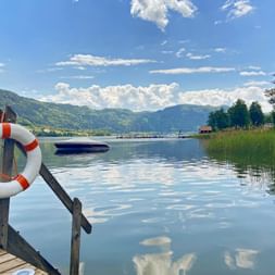 Wooden dock on Ossiachersee with white life ring, calm lake reflecting clouds, boat in distance, green mountains and reeds along shore.