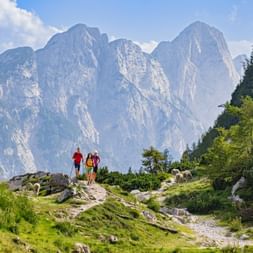 Three hikers on rocky trail at Vršič Pass in Slovenia with sheep grazing nearby. Dramatic mountain peaks rise in background under blue sky.