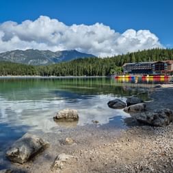 Felsiges Ufer des Eibsees mit klarem türkisfarbenem Wasser, bunten Booten und Gebäuden. Dichter Wald und Zugspitze unter blauem Himmel.