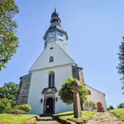 Weiße Kirche mit dunklem Turm und Uhr in Reinhardtsdorf. Steinkreuz im Vordergrund, grüner Rasen und Bäume rahmen das Gebäude unter blauem Himmel.