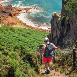 Wanderer mit Rucksack und roter Shorts auf Küstenpfad mit Blick auf türkisfarbenes Meer und Felsklippen mit grüner Vegetation auf den Kanalinseln.