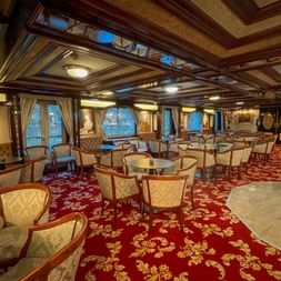 Luxurious ship lounge with ornate ceiling, red patterned carpet, round tables with cream chairs, and large windows showing river views.