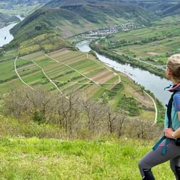 Wanderin mit blauem Rucksack sitzt am Hang mit Blick auf die Moselschleife mit Weinbergen, grünen Feldern und Dörfern in Deutschland.