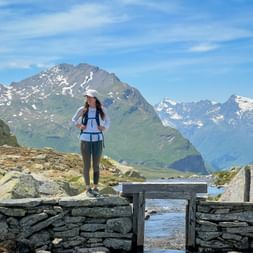 Female hiker on wooden bridge over mountain lake Surettasee with snow-capped Alpine peaks in background under blue sky on Via Spluga trail.
