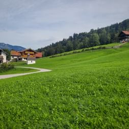 Grüne Wiese mit gewundenem Weg zu traditionellen Alpenhäusern in Graseck. Bewaldete Hügel und Berge im Hintergrund.