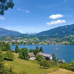 Panoramic view of Millstättersee lake with Seeboden town on the opposite shore, surrounded by green mountains and forests under blue sky.