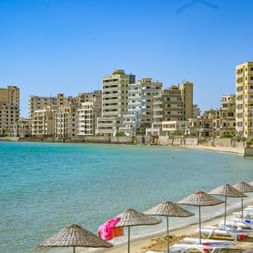 Strand mit Sonnenschirmen und Liegen vor verlassenen Apartmentgebäuden der Geisterstadt Varosha. Kristallklares türkisfarbenes Wasser unter blauem Himmel.