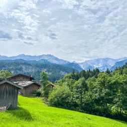 Traditionelle Holzhütten auf grüner Wiese mit Alpengipfeln im Hintergrund. Dichte Wälder bedecken die Hänge unter bewölktem Himmel bei Garmisch.