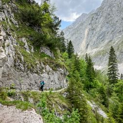 Wanderin in blauer Jacke auf schmalem Bergpfad entlang Felswand am Höllentalklamm, umgeben von Nadelbäumen und steilen Bergwänden.