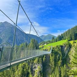 Metallene Hängebrücke über einer bewaldeten Schlucht in Holzgau mit grünen Almwiesen, Bergen und blauem Himmel im Hintergrund.