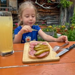Young girl in blue dress eating traditional alpine snack with salami and pickles on wooden board at Kammeregg Alm. Rustic cabin and flowers visible.