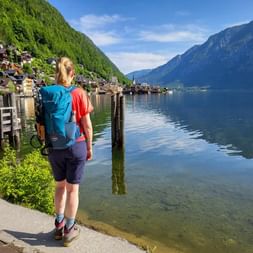 Female hiker with blue backpack viewing Hallstatt village across calm lake waters, surrounded by steep forested mountains under blue sky.