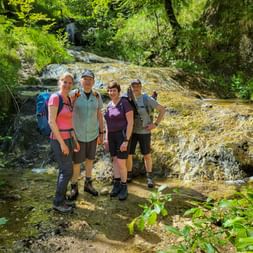 Four hikers with backpacks standing in a stream before a cascading waterfall surrounded by lush green forest.