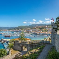 View from Baiona's stone fortress overlooking the marina with sailboats, coastal town, and green hills under blue sky.