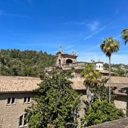 Stone buildings of Lluc Monastery with terracotta roofs, palm trees, and mountains under blue sky in Mallorca on the GR221 hiking trail.