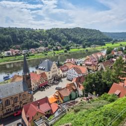 Luftaufnahme von Stadt Wehlen mit Kirche, Marktplatz und rotgedeckten Häusern entlang der Elbe, umgeben von grünen Hügeln.