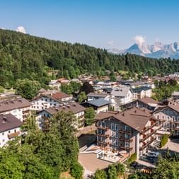 Aerial view of Kitzbühel village with traditional Alpine buildings, surrounded by dense forests and mountain peaks in the background under blue sky.