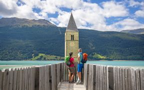 Wanderer blicken auf den türkisen Reschensee mit seinem versuckenen Kirchturm