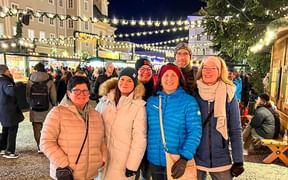 Sechs Teammitglieder in Winterkleidung lächeln auf einem Christkindlmarkt mit festlicher Beleuchtung und Marktständen am Abend.