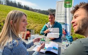 Three people at outdoor table with Eurohike banner, discussing hiking brochures in sunny meadow with forest hills in background.