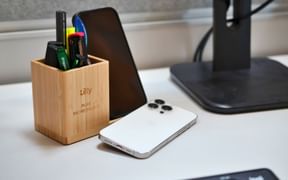Weißer Schreibtisch mit Bambus-Stiftehalter, zwei Smartphones und Tastatur. Der Stiftehalter enthält Stifte und Marker. Ein schwarzer Monitorständer ist sichtbar.