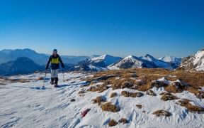 Wanderin mit Stöcken auf verschneitem Bergweg der Postalm Moosalmrunde mit schneebedeckten Gipfeln und blauem Himmel im Hintergrund.