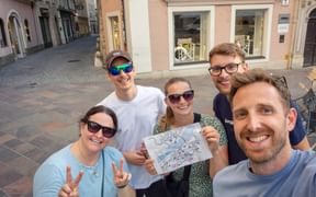 Five smiling team members posing with a city map on a cobblestone square. Historic buildings with colorful facades are visible in the background.