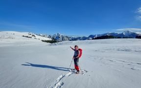 Wanderer mit rotem Rucksack und Stöcken winkt auf verschneiter Postalm. Berggipfel unter klarem blauen Himmel im Hintergrund sichtbar.