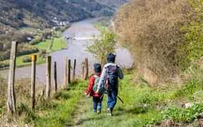 Two hikers with backpacks walking down a grassy hillside path with wooden fence posts, overlooking the Moselle River valley below.