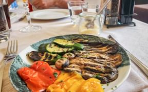 Plate of grilled vegetables including zucchini, eggplant, bell peppers, and mushrooms on a restaurant table with wine glasses and condiments.