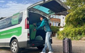 Man loading luggage into white and green Eurohike van with open rear doors. Black suitcase stands nearby on pavement with house in background.