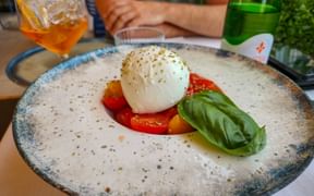Caprese salad with burrata cheese, sliced tomatoes, and fresh basil on a speckled plate in Bardolino, Lake Garda. Drinks visible in background.