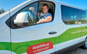 Lächelnder Fahrer in weiß-grünem Eurohike-Transporter mit Gepäcktransport-Beschriftung. Der Transporter zeigt eurohike.at Logo und Telefonnummer.