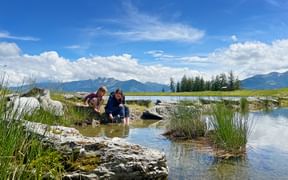 Two children crouching at the edge of a mountain reservoir in Pinzgau, exploring shallow water among rocks and grass under blue sky.
