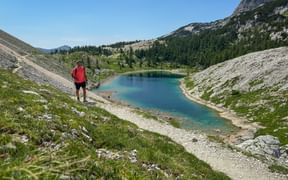 Wanderer in roter Jacke mit Blick auf türkisen Bergsee im Sieben-Seen-Tal, Julische Alpen, mit felsigen Hängen und bewaldeten Gipfeln.