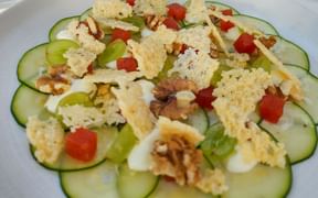 Zucchini carpaccio arranged on a white plate with crumbled cheese, walnuts, red bell pepper pieces, and green grapes.