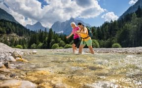Two hikers with backpacks crossing a shallow mountain stream at Jasna Lake, surrounded by forested mountains and dramatic alpine peaks.