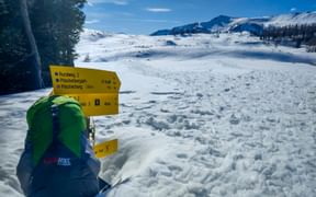 Gelber Wegweiser mit Rucksack in verschneiter Landschaft auf der Postalm Pitschenberg. Schneebedeckte Berge und blauer Himmel im Hintergrund.