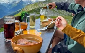 Two hikers enjoying soup in wooden bowls at mountain hut table with drinks, overlooking Alpine peaks in Lechtal.