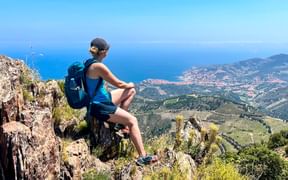 Female hiker with backpack sitting on rocky outcrop overlooking Collioure coastline with blue Mediterranean Sea and green hills.