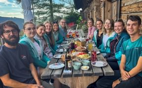 Large group of Eurohike team members sitting around wooden table on terrace of traditional alpine hut, sharing meal with mountain view.