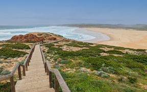 Holzsteg führt zum Praia da Bordeira Strand mit weißem Sand, Meereswellen und grüner Küstenvegetation unter blauem Himmel.
