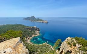 Rocky viewpoint on Mallorca's GR221 trail showing turquoise Mediterranean bay and forested island. Clear blue sky above calm waters.