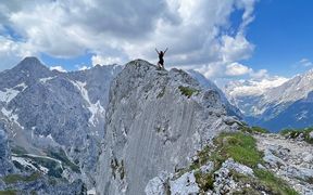 Wanderin steht auf einem Felsen inmitten der Bergwelt und streckt die Arme in Richtung Himmel