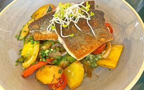 Crispy pan-fried char fillet topped with sprouts, served on a bed of colorful vegetables including yellow potatoes, bell peppers, and herbs.
