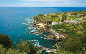 Luftaufnahme des Dorfes Caloura auf den Azoren mit weißen Gebäuden, üppigem Grün, felsiger Küste und türkisfarbenem Atlantikwasser.