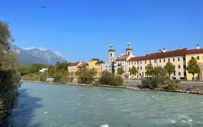 Türkisfarbener Inn fließt durch Innsbruck mit historischen Gebäuden und Kirchtürmen am Flussufer, Berge im Hintergrund.