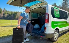 Mann lädt schwarzen Koffer in weißen Eurohike Gepäcktransport-Van mit grüner Beschriftung, geparkt auf Schotterweg neben grüner Wiese und Wald.